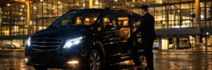 Conductor profesional uniformado abre la puerta de un monovolumen Mercedes premium para los pasajeros en la terminal del Aeropuerto de Berlín Brandenburg (BER) por la noche